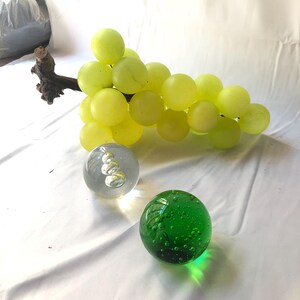 Può includere: Un grappolo di uva di vetro verde con un tralcio di legno, una sfera di vetro trasparente con bolle e una sfera di vetro verde con bolle.