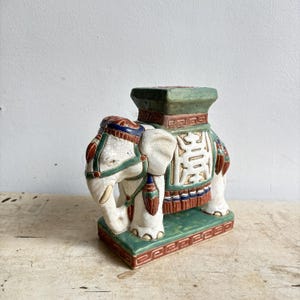 Può includere: Statuetta di elefante in ceramica nei colori verde, rosso e bianco. L'elefante ha una sella decorativa e una parte superiore piatta. La statuetta è su una base rettangolare con un bordo dettagliato. La proboscide dell'elefante è curva verso il basso.