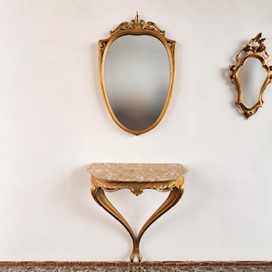 Può includere: Uno specchio da parete dorato con una cornice ornata a forma di scudo e uno specchio più piccolo di stile simile alla sua destra. Sotto, una console da parete presenta un piano in marmo beige e una base dorata curva con due gambe.
