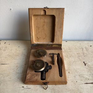 Könnte beinhalten: Eine Holzkiste mit einem Scharnierdeckel, die ein Set aus drei Metallwerkzeugen enthält. Die Werkzeuge umfassen einen runden Fräser mit einer Messingmitte, einen kleineren runden Fräser und einen Schraubenschlüssel aus Metall.