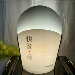 Può includere: Una lampada bianca rotonda illuminata con un manicotto di carta bianco e nero. Il manicotto ha il testo "快音感" e il logo Heineken.