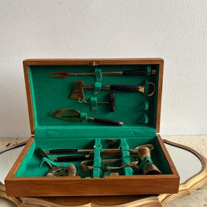 Op de afbeelding: Een vintage cocktail bar gereedschapsset in een houten kist. De set bevat diverse gereedschappen met messing en zwarte accenten, waaronder een kleine bijl, een taartschep en een jigger. De binnenkant van de kist is gevoerd met groen vilt.
