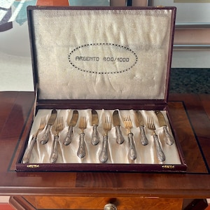 May include: A set of silver-toned flatware including forks and knives with ornate handles, arranged in a dark burgundy presentation case. The interior lid features a silver fabric lining with the text ARGENTO 800/1000 printed in a central oval.