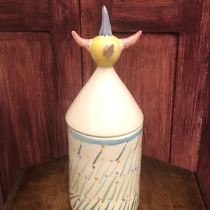 Può includere: Un barattolo in ceramica con un design di uccello fantasioso. L'uccello ha un corpo giallo, un cappello blu e ali rosa. Il barattolo è decorato con un motivo verde, giallo e blu.