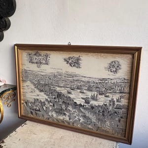 May include: A framed black and white illustration of London, England, with the River Thames running through the center. The illustration shows the city in the 17th century, with many buildings and ships. The frame is gold and has a simple design.