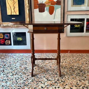 Puede incluir: Una mesa de madera marrón con alas abatibles que cuenta con un cajón central único con un pequeño pomo metálico redondo. La mesa tiene cuatro patas delgadas y torneadas conectadas por una barra transversal horizontal, ofreciendo una base estable.