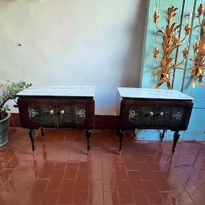 May include: Two antique bedside tables with marble tops. The tables have dark wood frames with decorative carvings and brass hardware. Each table has two doors and stands on four legs.