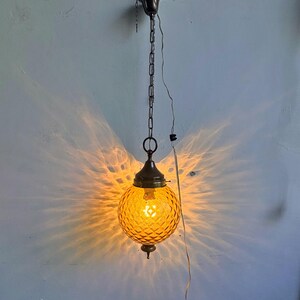Può includere: Una lampada a sospensione vintage con un globo in vetro ambrato strutturato. La lampada è sospesa da una catena e presenta un baldacchino e dettagli in metallo color bronzo. Il globo illuminato proietta un bagliore caldo e modellato sulla parete.
