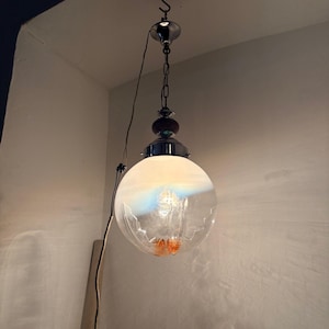 May include: A vintage pendant light fixture with a clear, spherical glass shade. The shade has a frosted top and a decorative orange element inside. The fixture includes a chrome chain and a dark brown and chrome accent.