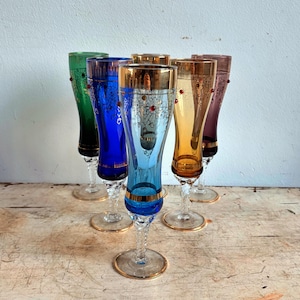 Può includere: Un gruppo di sei flûte da champagne vintage colorate. Ogni bicchiere presenta un colore diverso, tra cui verde, blu, ambra e viola, con bordi dorati e dettagli decorativi. Gli steli hanno un design a spirale.