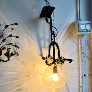 Può includere: Un lampadario a sospensione in ferro battuto nero con un paralume in vetro trasparente. La luce è appesa a una catena e ha una singola lampadina.