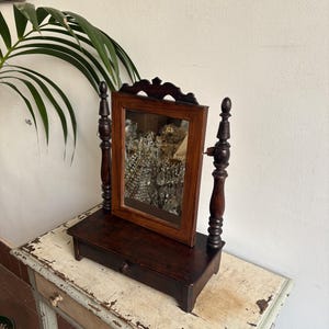 Peut inclure: Un miroir de coiffeuse antique marron foncé avec un cadre rectangulaire et des supports en bois décoratifs. Le miroir repose sur une petite base avec un tiroir. Le miroir reflète des bijoux. La coiffeuse est sur une table blanche vieillie.
