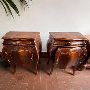 Puede incluir: Dos mesitas de noche antiguas de madera con acabado marrón oscuro. Cada mesa presenta un frente curvo, un solo cajón y herrajes de latón ornamentados. Las mesas tienen un diseño clásico y vintage con patas curvas e incrustaciones decorativas.