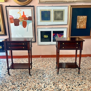 Peut inclure: Une paire de tables d'appoint en bois brun foncé assorties, chacune dotée de deux tiroirs superposés avec de petits boutons ronds en laiton, de pieds tournés fins et d'une étagère inférieure ouverte. Les tables reposent sur de petites roulettes.