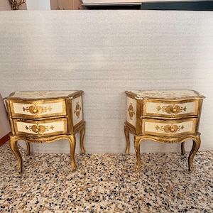 May include: A pair of bedside tables featuring two drawers each, finished in a cream and gold colour palette. The furniture pieces have curved cabriole legs and ornate gold-painted floral motifs on the drawer fronts and side panels.