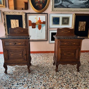 Op de afbeelding: Een paar bijpassende houten nachtkastjes met donkere marmeren bladen, een lade en een kastdeur. Elk meubelstuk is voorzien van decoratieve goudkleurige inlegpatronen op de fronten en rust op gesneden, gebogen poten.