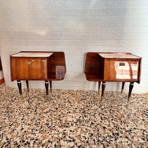 May include: A pair of matching bedside tables featuring a glossy dark wood finish, a white marble top, and a single drawer with a rectangular brass handle. Each table is supported by four tapered dark wood legs with brass-toned metal feet.