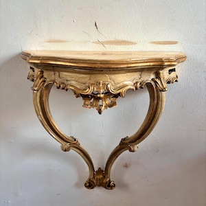 Op de afbeelding: Een wandconsole met een halfrond blad en een enkele gebogen steunpoot. Het meubelstuk is afgewerkt in een verweerde goudtint met gedetailleerd houtsnijwerk in krulvormen en florale motieven langs de rand en de voet.