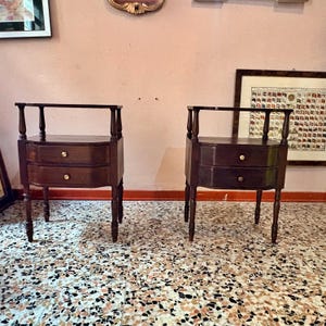 May include: Two antique wooden bedside tables. Each table has two drawers with brass knobs, supported by four slender legs. A small shelf is at the top, connected by four vertical posts. The tables are a dark brown colour.