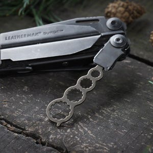 Peut inclure: Un outil multifonction Leatherman noir avec un accessoire de clé à molette argenté. L'outil est ouvert et la clé à molette est fixée sur le côté.