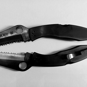 May include: Two black folding knives with serrated blades. The top knife has the word "POLICE" printed on the blade.