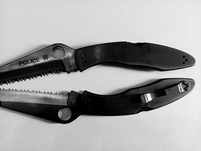 May include: Two black folding knives with serrated blades. The top knife has the word "POLICE" printed on the blade.