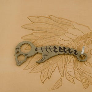 Könnte beinhalten: Ein silbernes Multitool mit einem Totenkopfdesign und einem Flaschenöffner. Das Werkzeug ist mit 3/8, 1/4 und 5/16 Zoll gekennzeichnet. Das Werkzeug befindet sich auf einem braunen Lederhintergrund mit einem indianischen Kopfschmuckdesign.