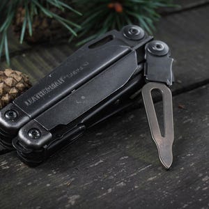 Könnte beinhalten: Ein schwarzes Multitool mit dem Text "Leatherman" auf der Seite. Das Werkzeug verfügt über eine Vielzahl von Klingen und Werkzeugen, darunter ein Messer, eine Säge und einen Schraubendreher. Das Werkzeug ist geschlossen und liegt auf einer Holzoberfläche.