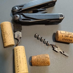 May include: A black multi-tool with the text "Leatherman SURGE" on the handle, a corkscrew, and four wine corks. One cork has the text "Varvaglione" and another has the text "L 288 N5 100096".