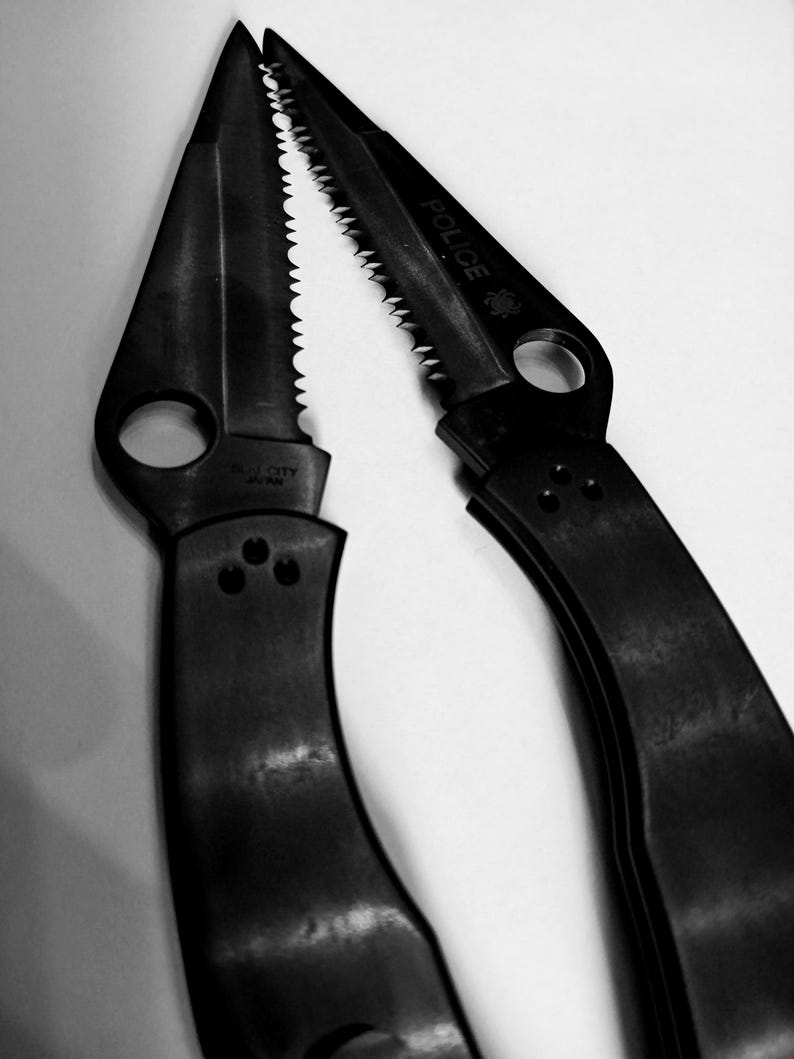 May include: A black folding knife with a serrated blade and a "POLICE" inscription. The knife has a black handle and a silver metal clip.