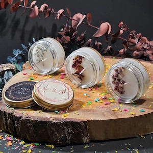 Puede incluir: Tres velas pequeñas en frasco de vidrio con cera blanca, cada una decorada con diferentes elementos botánicos que incluyen escamas plateadas, pétalos de rosa secos y flores moradas. Hay dos tapas de metal dorado, una con "OLIVIA & BAILEY" y "K & M".