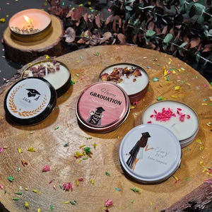 Op de afbeelding: Vier kaarsen met een afstudeerthema met verschillende ontwerpen. Eén kaars heeft een zwart en gouden ontwerp met het woord "Jack" en een afstudeerpet. Een andere kaars heeft een roze en gouden ontwerp met de tekst "Happy Graduation" en een afstudeerpet. Een derde kaars heeft een wit ontwerp met een afstudeerpet en de tekst "Class of 2020 Luna Simpson". De laatste kaars heeft een wit ontwerp met roze bloemblaadjes.