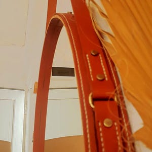 Puede incluir: Un espejo redondo con una correa de cuero marrón y herrajes de latón. El espejo está colgado de una pared con un fondo blanco.