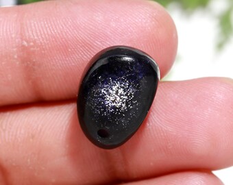 Könnte beinhalten: Ein dunkelblauer, ovaler Edelstein mit glitzerndem, sternenförmigem Aussehen. Der Stein hat ein kleines Loch, das durchbohrt wurde, was darauf hindeutet, dass es sich um einen Anhänger oder einen Glücksbringer handelt.