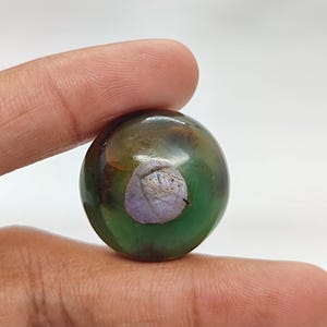 Może przedstawiać: Okrągły, zielono-brązowy kaboszon agatowy z wtrąceniem fioletowym i białym. Kaboszon jest gładki i wypolerowany.