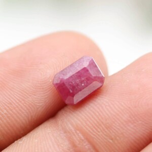 May include: A faceted, rectangular ruby gemstone. The gem is a deep pinkish-red color with a polished surface. The gemstone is held in a hand, with the background out of focus.