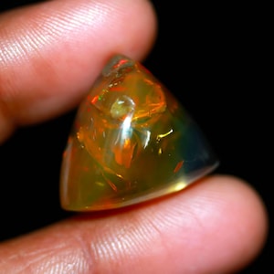 Puede incluir: Una gema de ópalo triangular pulida, translúcida, con una mezcla de tonos naranja, amarillo y verde, con destellos rojos internos. La gema se sostiene entre los dedos sobre un fondo negro.