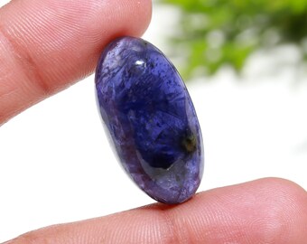 May include: A deep purple oval cabochon gemstone with a shimmering, iridescent finish. The stone is held between two fingers, showcasing its size and shape.