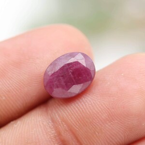 May include: A faceted, oval-shaped ruby gemstone. The ruby is a deep, saturated red color with multiple facets that catch the light. The gemstone is held between fingers, showcasing its size and shape.