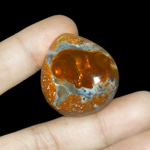 May include: A teardrop-shaped, orange and white opal cabochon with a swirling pattern. The opal is set in a finger, showing its size and shape.