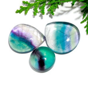 Könnte beinhalten: Drei Fluoritsteine in Grün-, Blau- und Violetttönen. Die Cabochons sind poliert und haben eine glatte, glänzende Oberfläche.