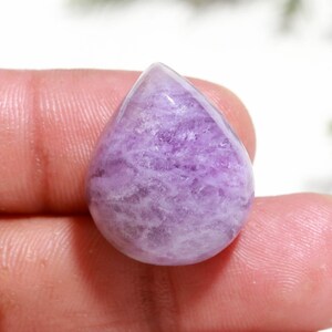 May include: A teardrop-shaped cabochon gemstone featuring a translucent light purple color with a textured, crystalline internal pattern. The stone has a smooth, polished surface and a rounded, symmetrical shape that tapers to a point at the top.