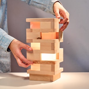 Può includere: Una lampada in legno realizzata con blocchi impilati con una luce che brilla attraverso gli spazi. La lampada è su una superficie bianca.