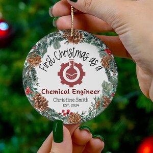 Puede incluir: Un adorno navideño redondo con un diseño de corona verde, blanca y marrón. El adorno presenta el texto "First Christmas as a Chemical Engineer" con un nombre personalizado y un año.