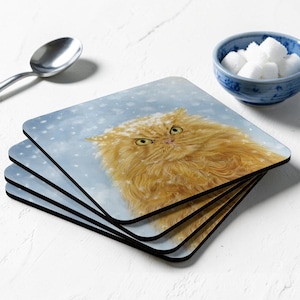 May include: A stack of four square coasters featuring a digital illustration of a fluffy orange cat with yellow eyes, set against a light blue background with white snowflakes. The coasters have rounded corners and a black edge finish.