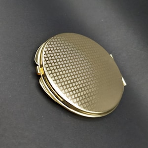 May include: A gold-colored, round compact mirror with a textured, repeating pattern on the top. The compact has a hinged opening and a shiny, metallic finish. The background is a dark gray.