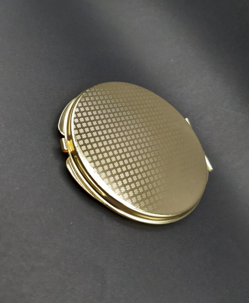 May include: A gold-colored, round compact mirror with a textured, repeating pattern on the top. The compact has a hinged opening and a shiny, metallic finish. The background is a dark gray.