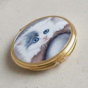 White and grey fluffy kitten with blue eyes hand drawn artwork, printed onto gold metal compact mirror with classic clasp opening.