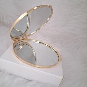 May include: A gold-colored, oval-shaped compact mirror, open to reveal two reflective surfaces. The mirror is resting on a white box against a soft, white background. The compact has a hinged design.
