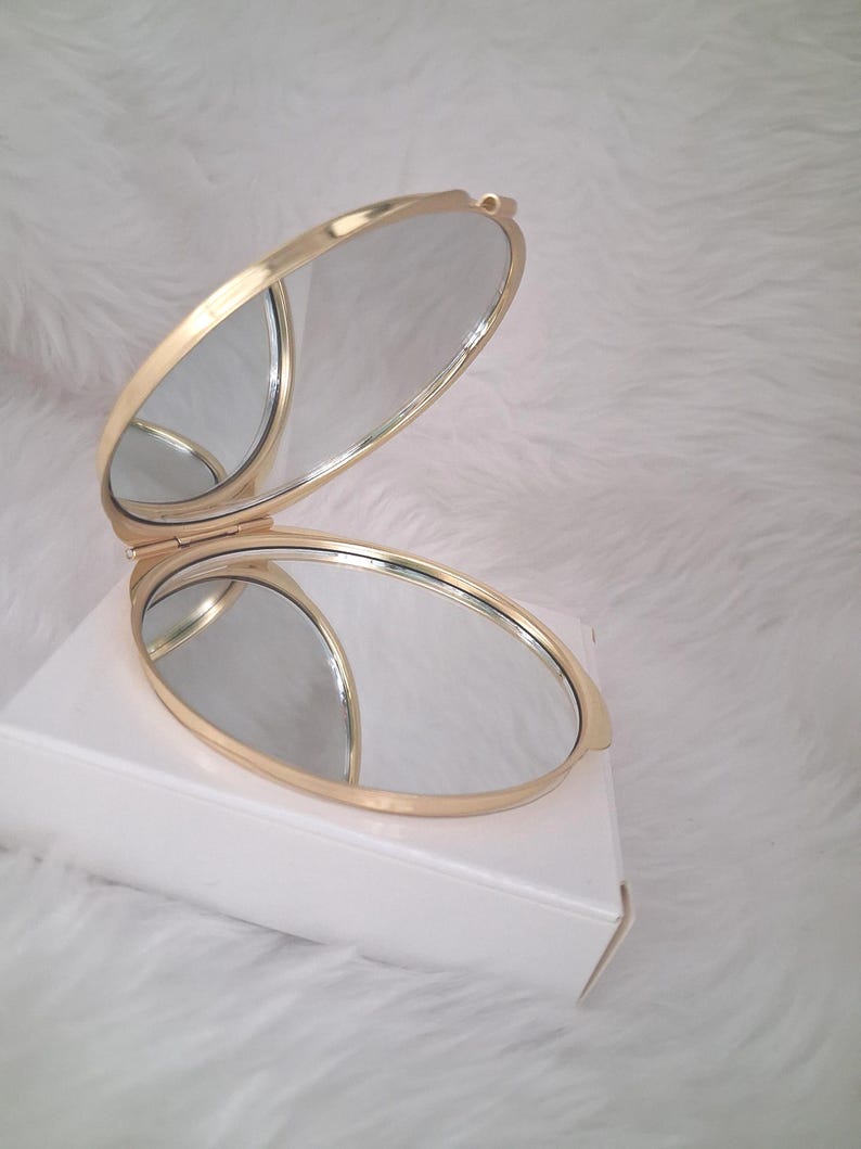 May include: A gold-colored, oval-shaped compact mirror, open to reveal two reflective surfaces. The mirror is resting on a white box against a soft, white background. The compact has a hinged design.